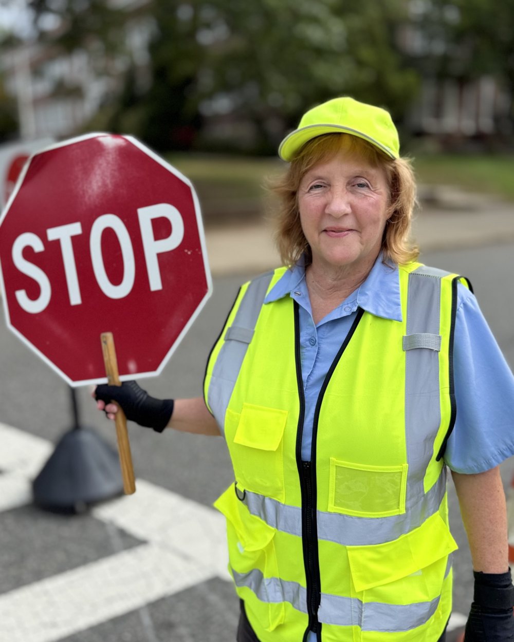 Eileen, in a yellow safety vest and cap, holding a red STOP sign at the crosswalk